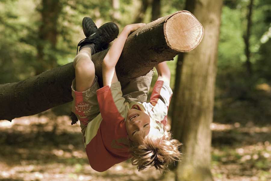 Een kind hangt aan ondersteboven aan een boomstam in het speelbos